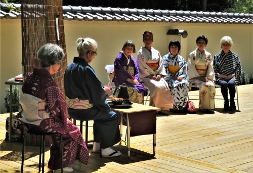 Jim sits at a small tea table with a line of 5 people sitting in the background on a large wooden deck in the Japanese garden